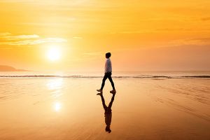 A person walks along the beach as the sun sets behind the ocean.